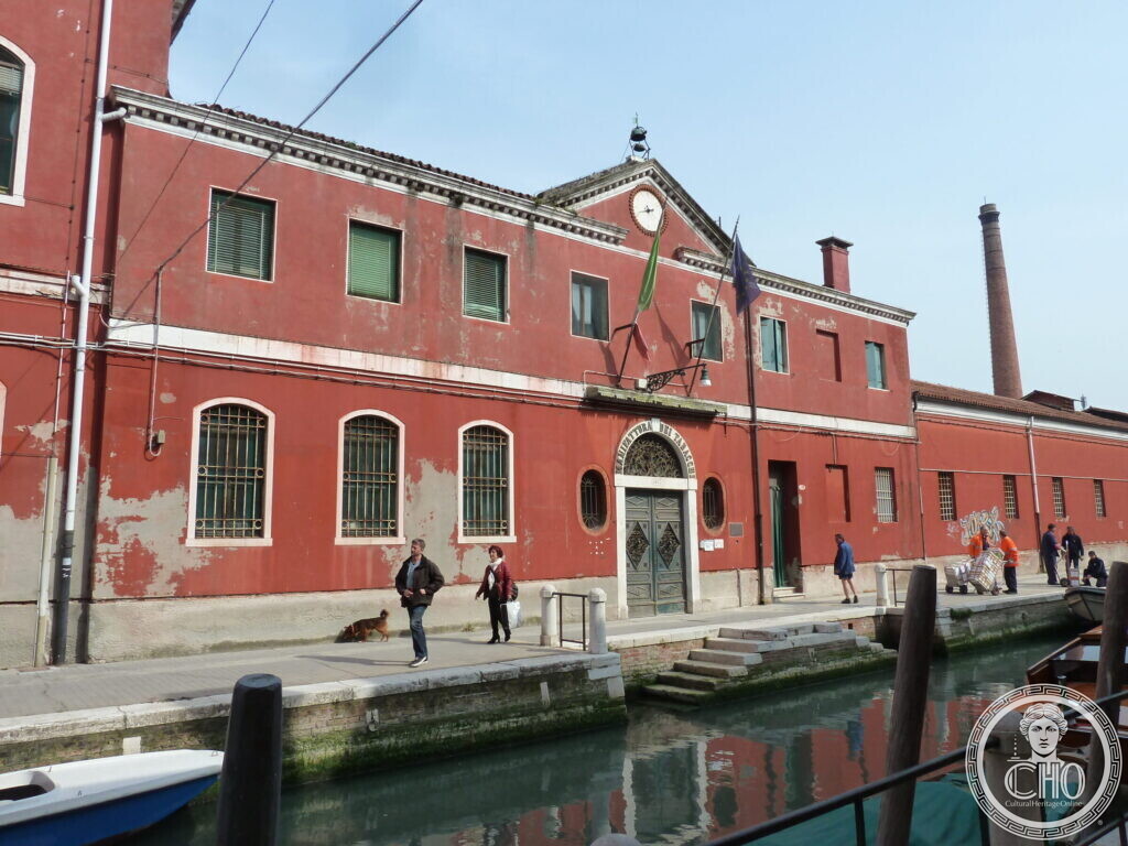 The red factory front running along a canal-side walkway with people walking, a tall brick chimney rising behind