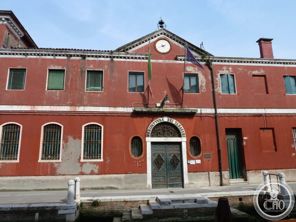 A red plastered two-storey factory front with a clock in the central pediment and the words Manifattura dei Tabacchi over an arched green door