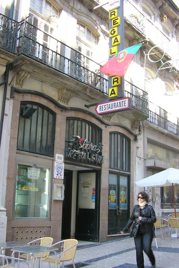 The front of the A Regaleira restaurant on Rua do Bonjardim in 2007: a ground floor clad in polished granite with three round-arched openings under a wavy cornice, neon lettering over the door, a vertical yellow sign reading Regaleira, and wrought-iron balconies on carved stone consoles above
