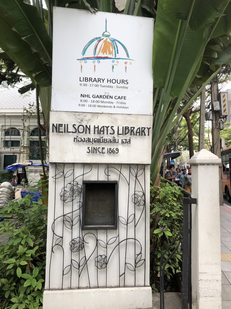 The marble gatepost of the Neilson Hays Library, lettered NEILSON HAYS LIBRARY in English and Thai with SINCE 1869 below, over a wrought-iron panel of roses, with a board giving the opening hours