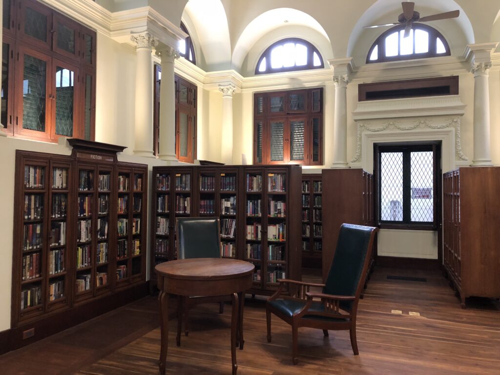 The reading room of the Neilson Hays Library: barrel vaults on Corinthian columns, arched clerestory windows, glazed teak bookcases lining the walls, a round table and two green leather armchairs on a teak floor