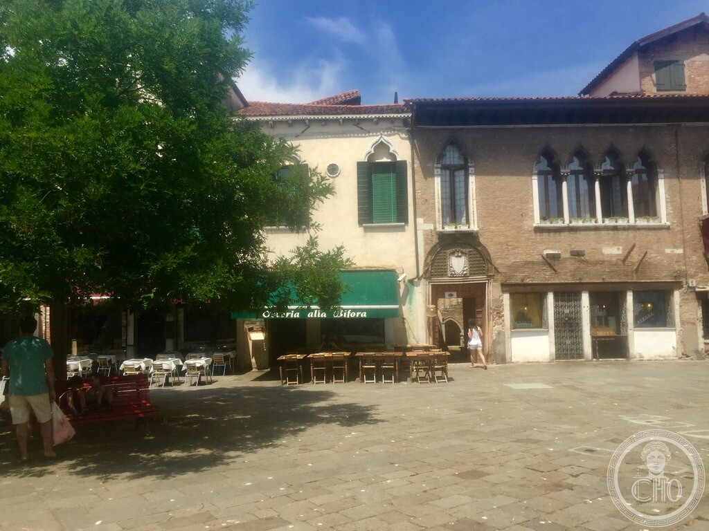 Wide perspective across Campo Santa Margherita showing Osteria Alla Bifora with outdoor seating under red and green awnings