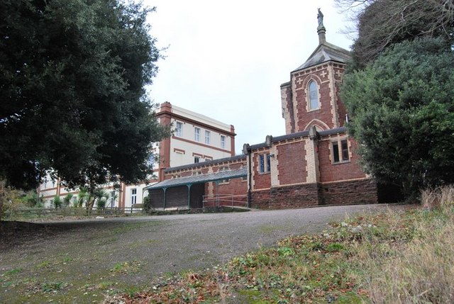 Former Marist monastery at Paignton: the red chapel end crowned by a statue, a lower range with a veranda and a cream three-storey house, framed by large evergreen trees