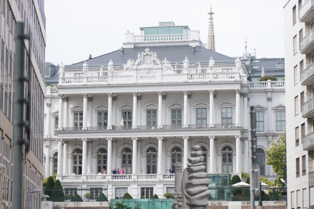 White late-classicist palace front with two tiers of free-standing columns, a balustrade of statues, a crowned coat of arms and the date MDCCCLXIV, raised above a brick bastion wall, Vienna