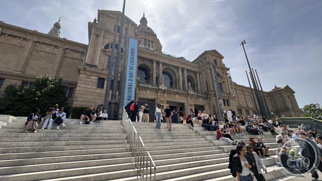 Visitors sitting on the wide stone steps leading up to the entrance of the Palau Nacional, a banner hanging on the facade
