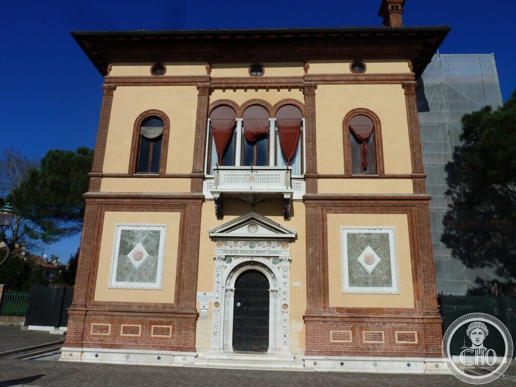 A small two-storey palace in brick and cream plaster with arched windows, a loggia with red curtains and a carved white stone portal
