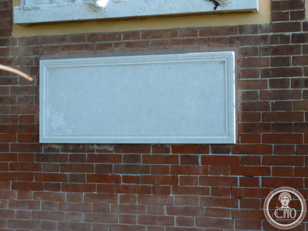 A plain rectangular marble tablet framed in white stone set into a red brick wall, the edge of a relief above
