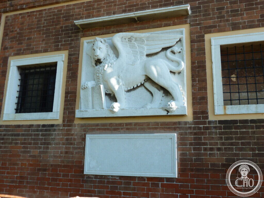 A white stone relief of the winged lion of Saint Mark set into a brick wall between two barred windows