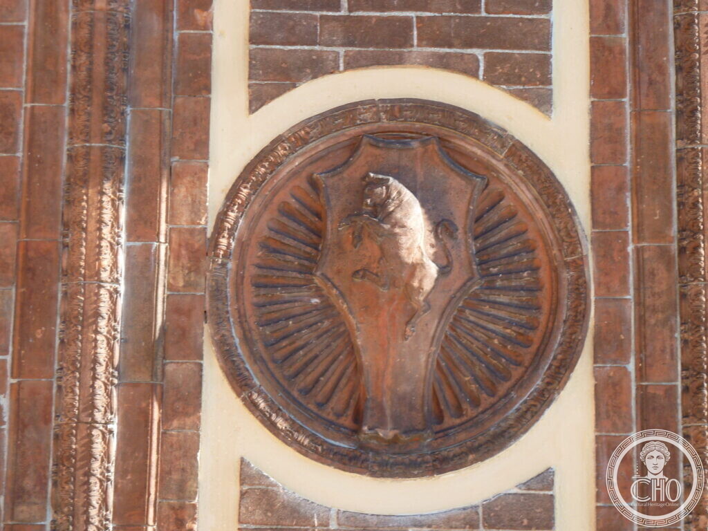 A round terracotta relief with a shield bearing a rampant lion against radiating rays, set in a brick wall
