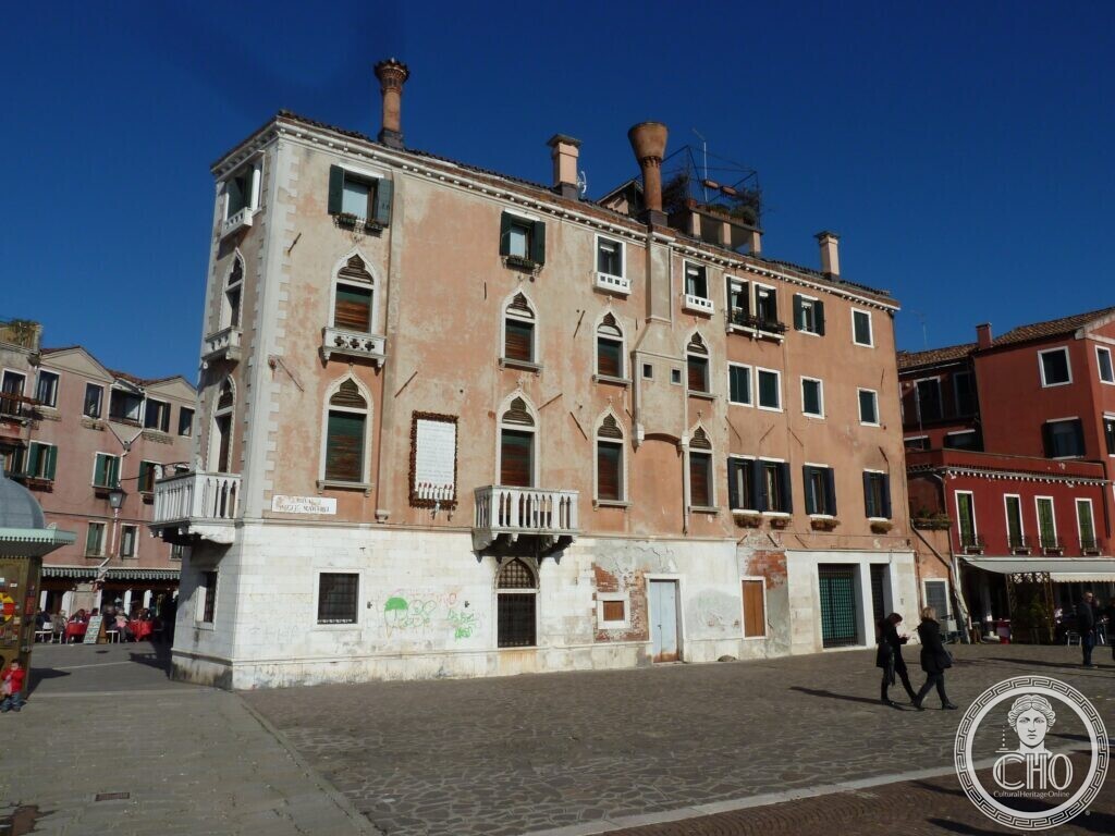 A tall plastered palace with pointed Gothic windows and small balconies on a street corner, tall chimneys on the roof
