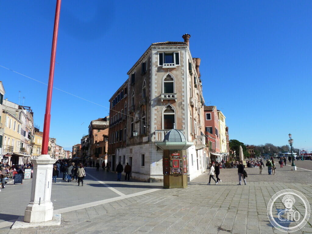 A narrow Gothic palace on a street corner with a kiosk in front, a wide waterfront promenade with people on the left