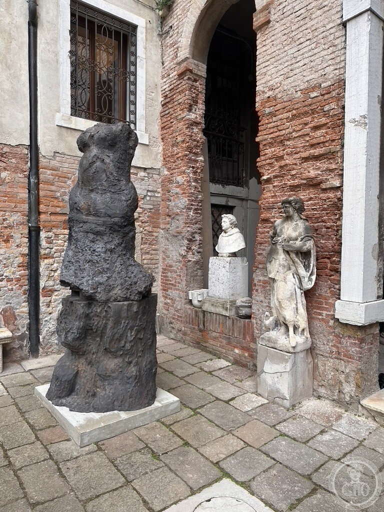 A dark contemporary sculpture standing beside a niche with an antique marble bust in the brick courtyard of Palazzo Contarini degli Scrigni e Corfù