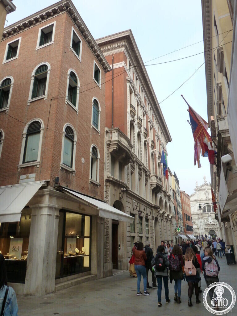A busy shopping street with a brick corner house in front and a taller pale stone palace with flags and a carved cornice beyond it