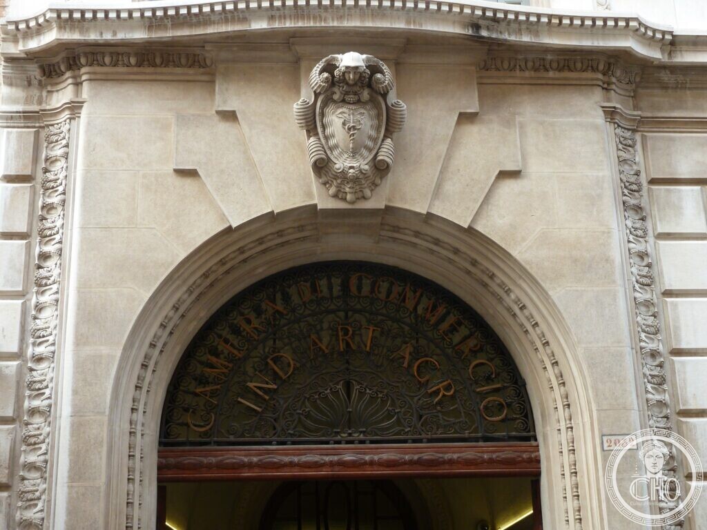 A carved stone arched doorway with a cartouche above and a wrought-iron lunette reading Camera di Commercio Ind Art Agr, house number 2032 on the right
