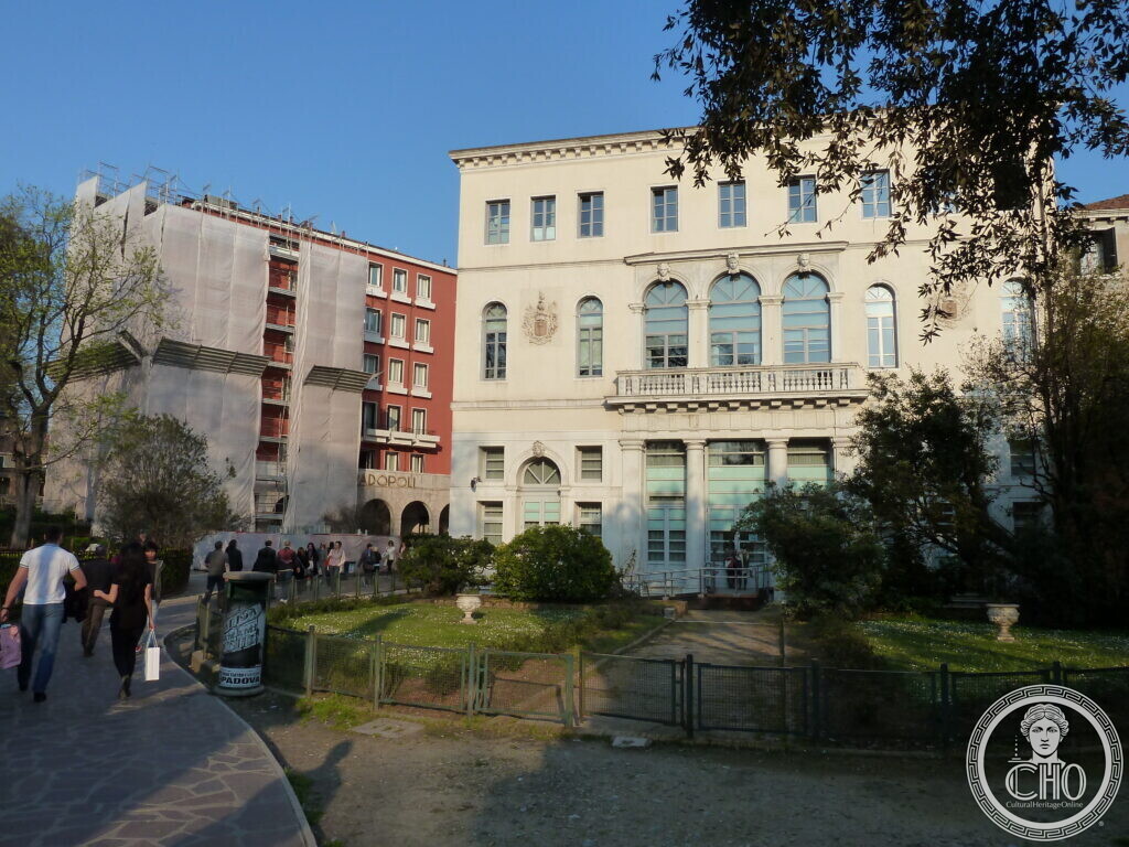 Palazzo Foresti Papadopoli seen at an angle from the Papadopoli gardens in Venice, the red block of the Hotel Papadopoli under scaffolding to its left and people walking along the garden path