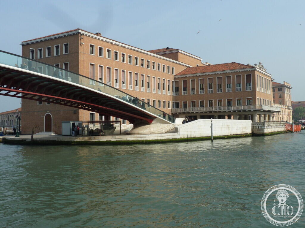 A long red brick office building with a loggia on the Grand Canal, the glass and steel arch of the Ponte della Costituzione landing beside it