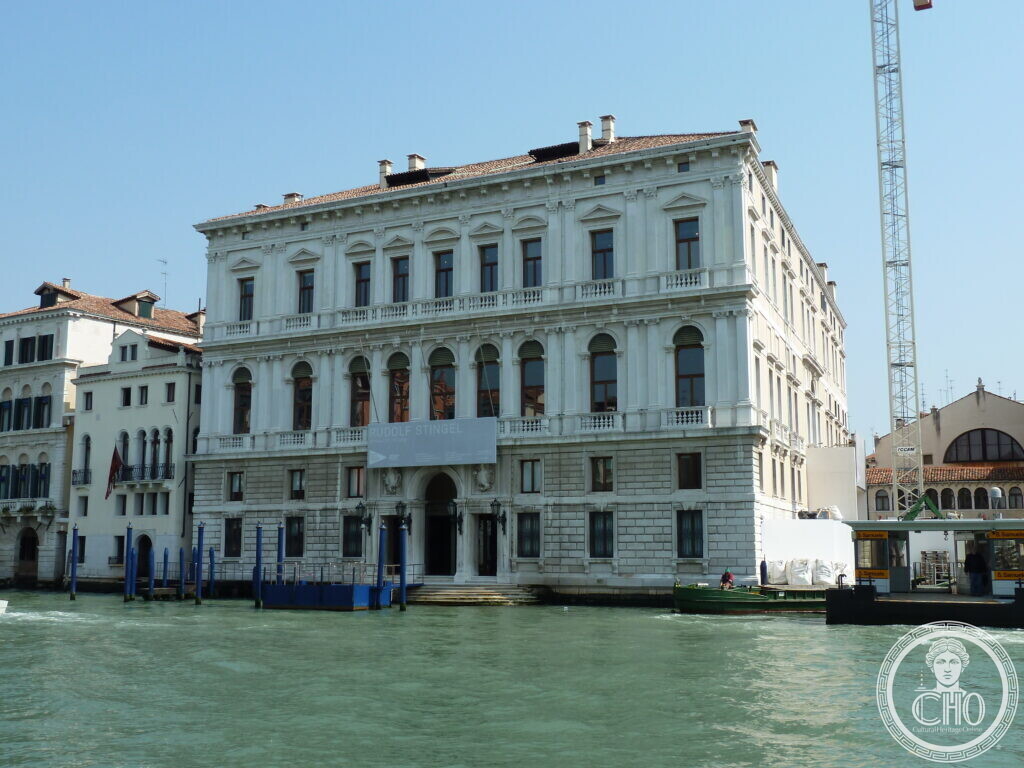 The white stone facade of Palazzo Grassi seen from the Grand Canal in Venice, with a banner for the Rudolf Stingel exhibition hung between the first-floor windows