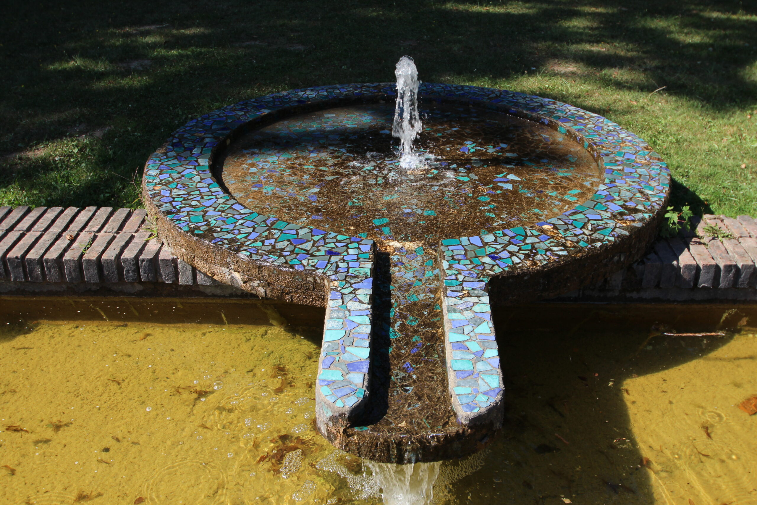 A circular basin of exposed-aggregate concrete edged with iridescent mosaic, water falling into a channel
