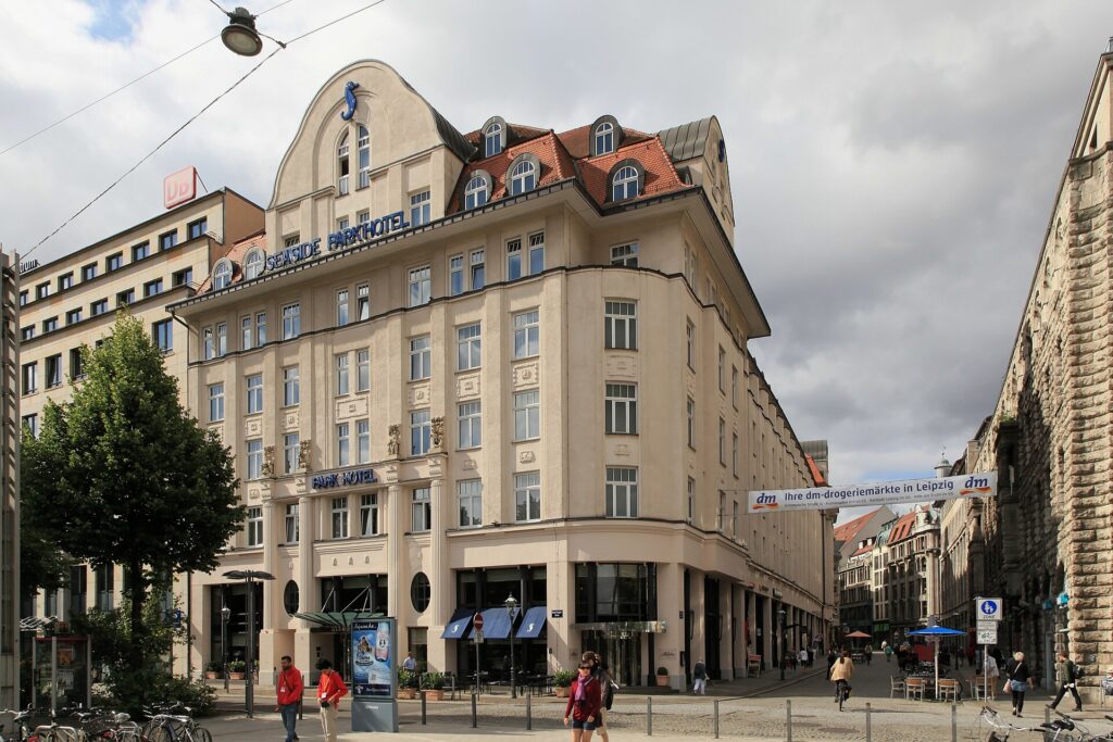 Corner building of the Park Hotel in Leipzig, pale rendered facade with pilasters, a tall curved gable, red-tiled mansard with round dormers, and oval windows beside the entrance on the ground floor