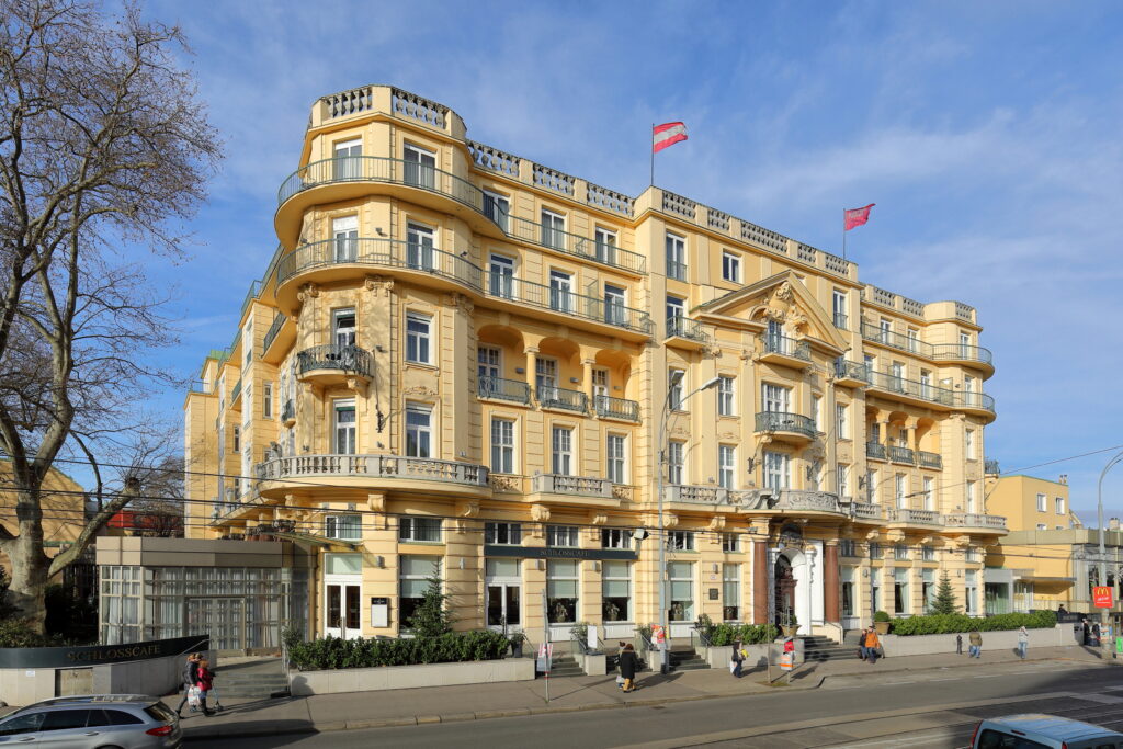 Large cream-yellow hotel of 1907 with rounded corner bays, curved balconies, a balustraded roofline and a carved gable, Hietzinger Hauptstraße, Vienna