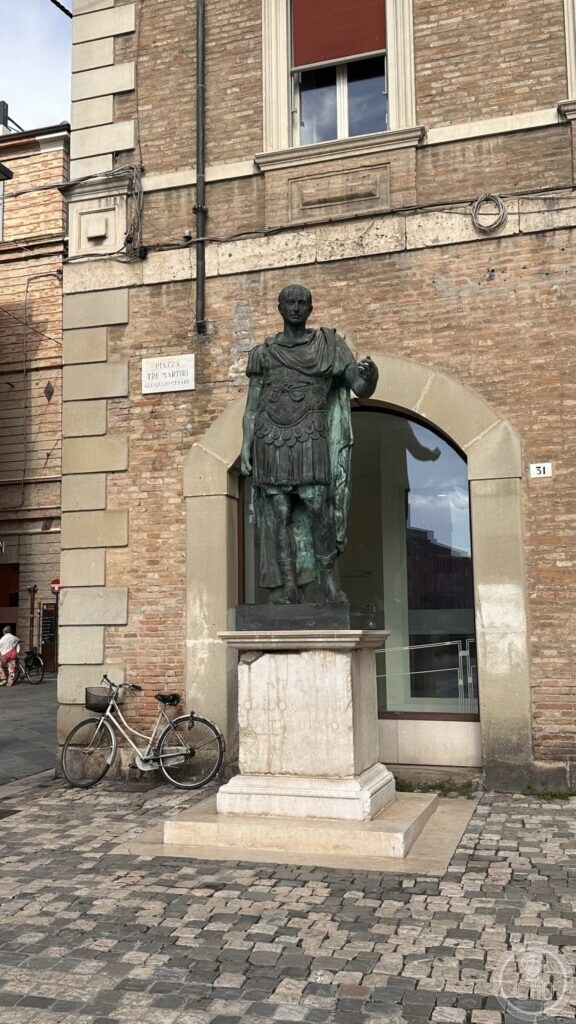 Bronze statue of Julius Caesar in armour on a stone pedestal against a brick wall, with the street sign above