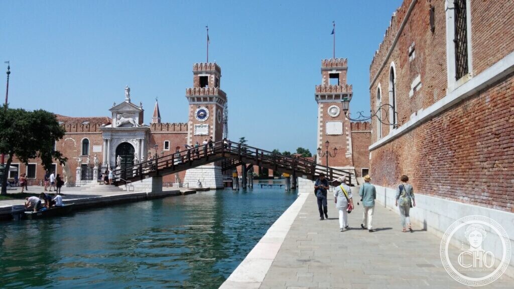 A wooden footbridge crossing a canal towards the Arsenal gate and two crenellated brick towers, people walking on the stone quay