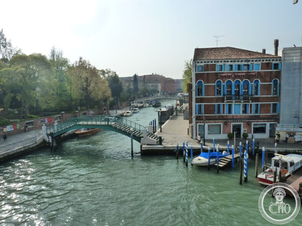 A green iron footbridge with stepped ramps crossing a canal, trees along one bank and a red hotel building with blue shutters on the corner