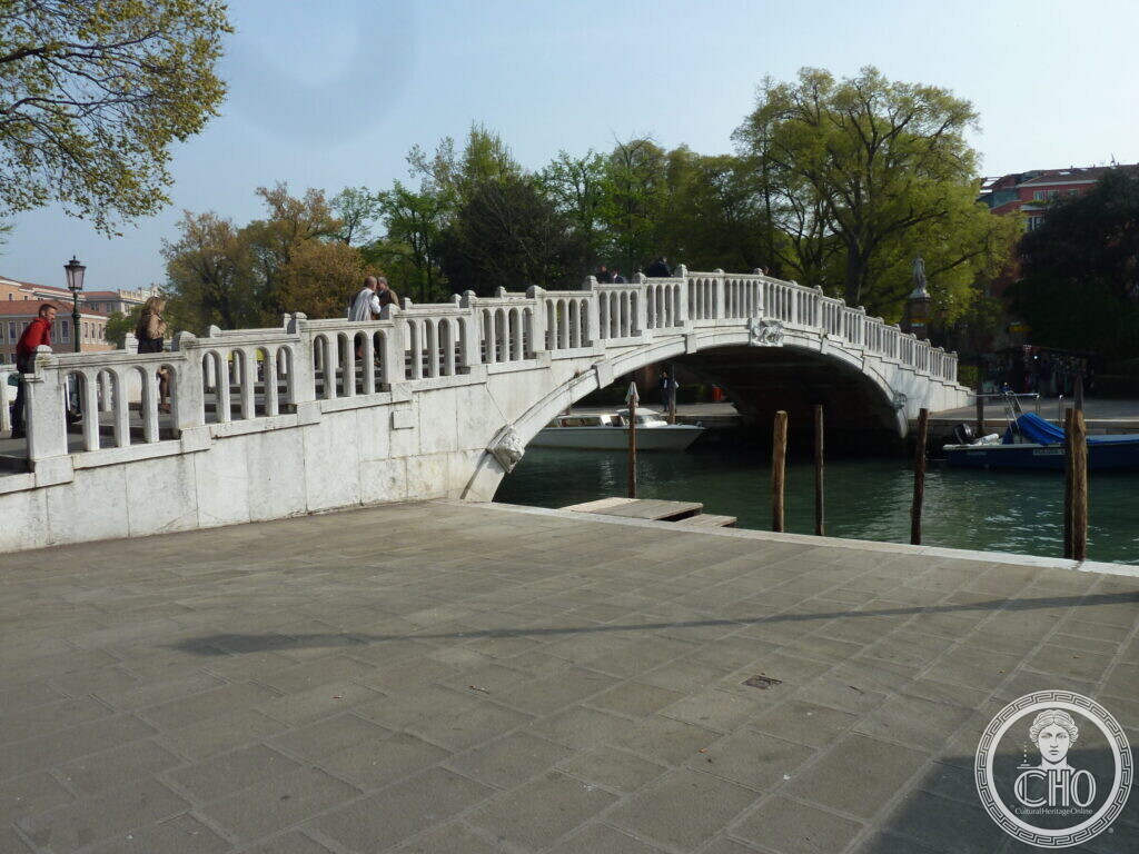 A single white stone arch bridge with a balustrade crossing a canal, trees in spring leaf behind it