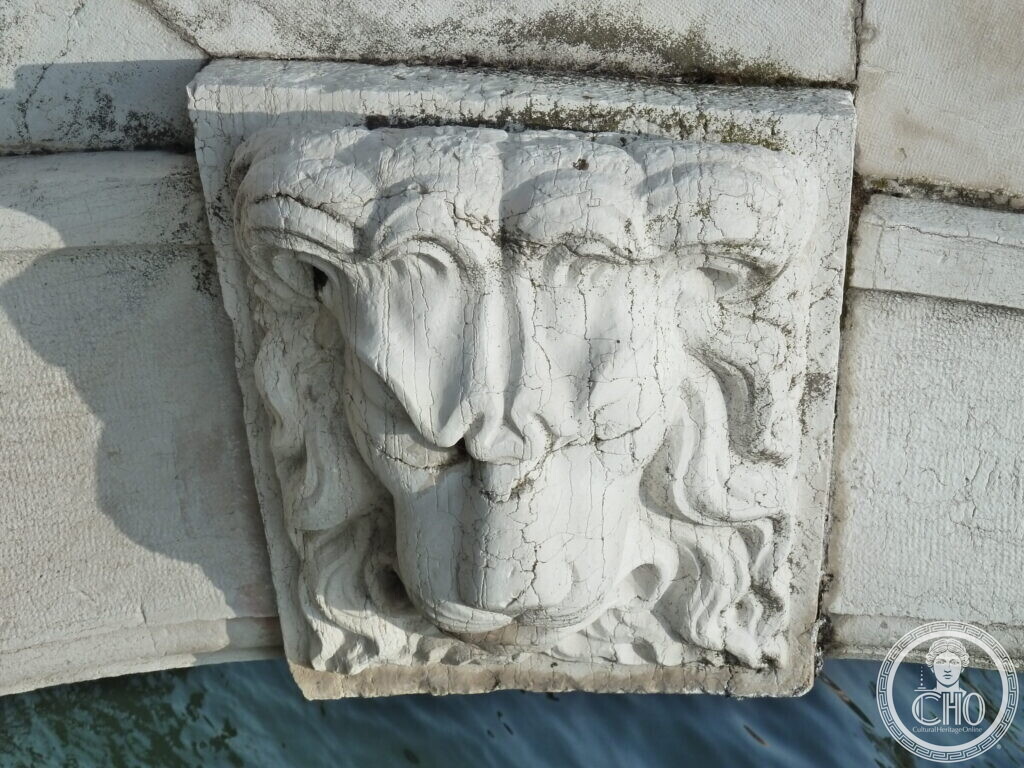 A close view of a weathered stone lion mask on the keystone of a bridge arch, just above the water