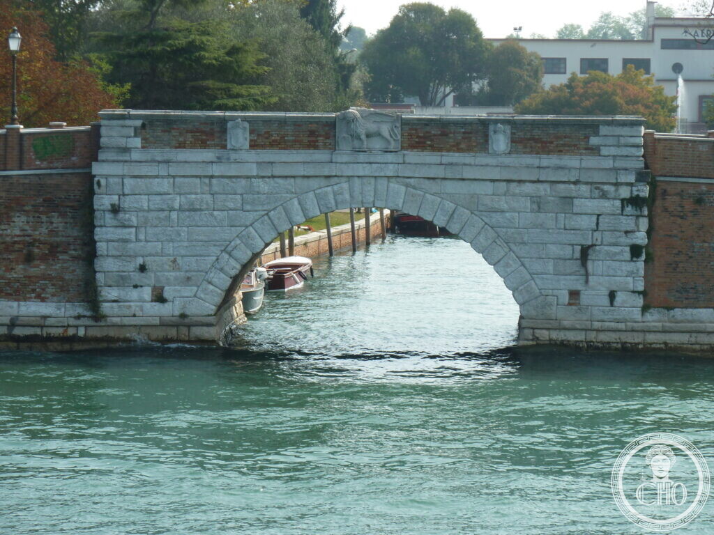 A single-arched stone bridge over a canal with a brick parapet, a carved winged lion and two shields in the middle, boats moored under the arch