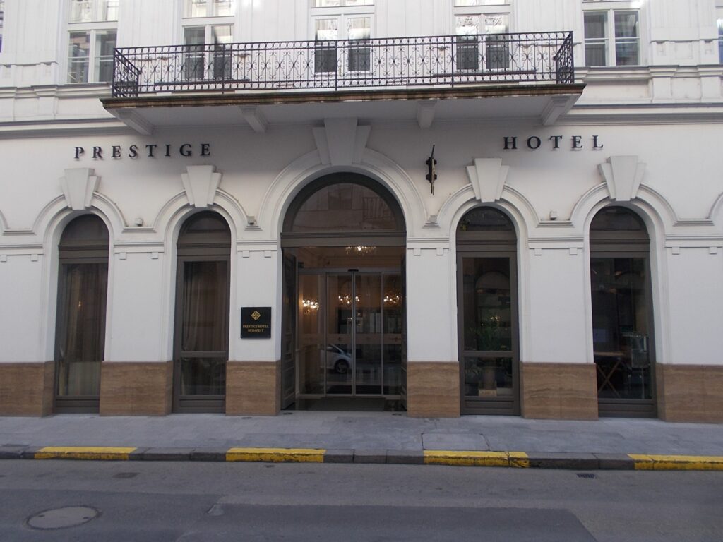 Ground floor and first-floor balcony of Vigyázó Ferenc utca 5, Budapest: a row of round-arched openings with a central glazed entrance, the lettering Prestige Hotel, and a wrought-iron balcony on brackets above