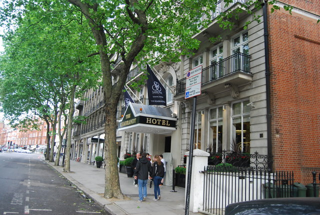 Stone front of the Rembrandt hotel at 11 Thurloe Place, London, with iron-railed balconies, a glass entrance canopy and flags, behind a line of street trees