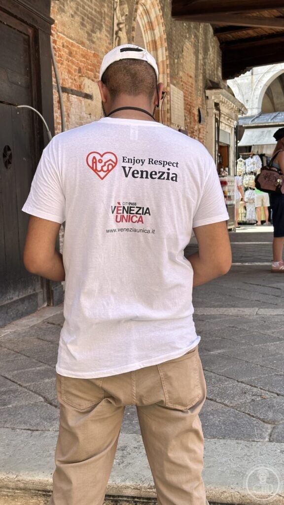 A man seen from behind wearing a white shirt printed Enjoy Respect Venezia, standing under the arcades near the stalls