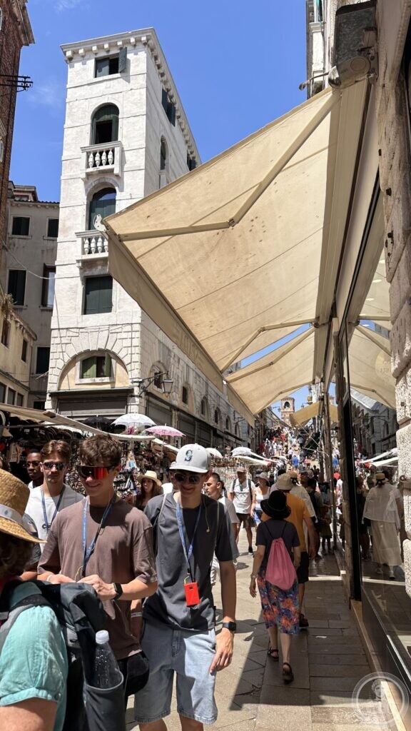 A narrow Venetian street packed with people under stall awnings, a bell tower visible at the far end