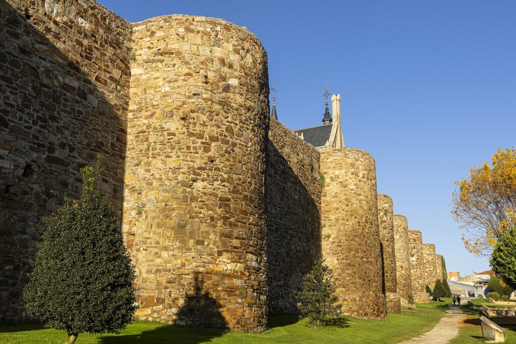 Stretch of the Roman walls of Astorga with a row of massive semicircular towers of rough stone beside a lawn and a path, the pinnacles of Gaudí's Episcopal Palace rising behind