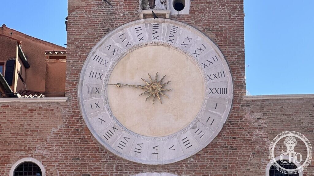 A large round stone clock face with Roman numerals from I to XXIIII and a gilded sun-shaped hand, set in a brick wall