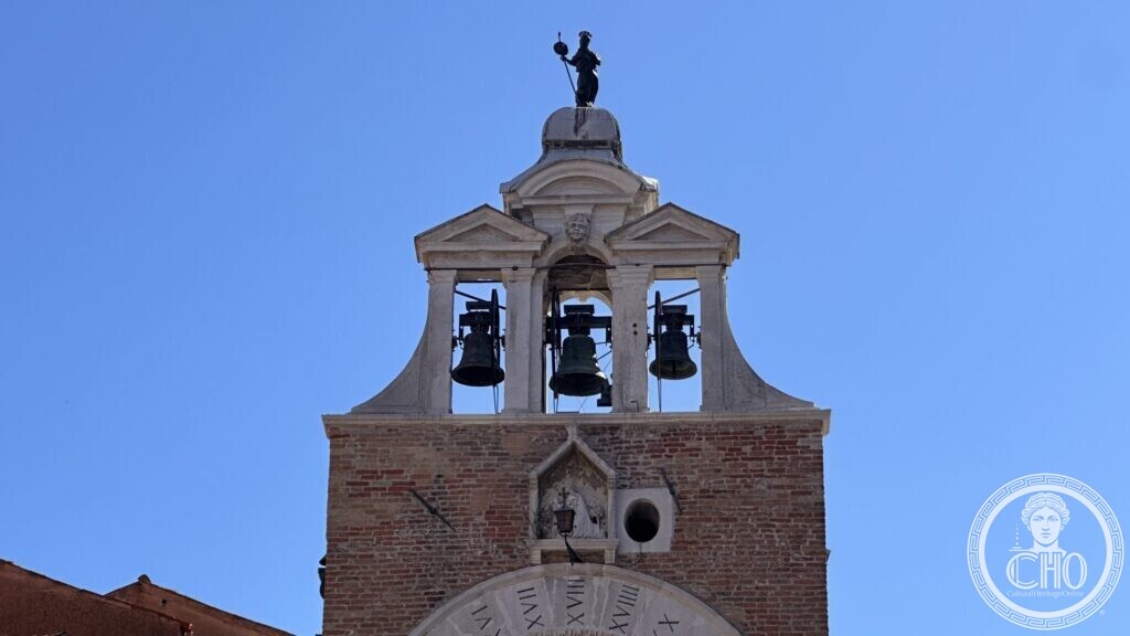 A brick and white stone bell gable with three bells in open arches, a small dome and a dark statue on top, against a blue sky