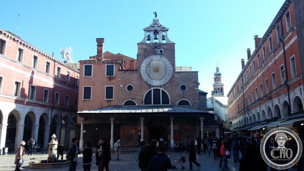 The front of San Giacomo di Rialto in Venice with its large single-hand clock, the five-column Gothic porch below and the bell gable with two bells above, seen across the busy campo