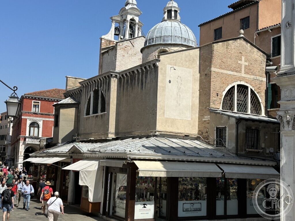 The lead dome and white bell gable of San Giacomo di Rialto rising behind the market shops, a sundial painted on the plastered wall below