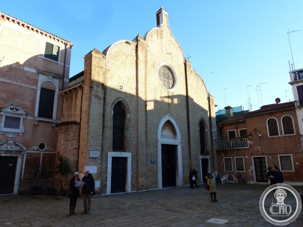 A tall brick church front divided into three parts with curved tops, a round window and two doorways, people standing in the paved square