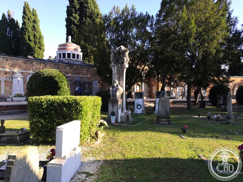 A lawn with scattered graves under tall cypresses at San Michele cemetery, a stone pillar with a bust and a sculpted group at the top in the middle