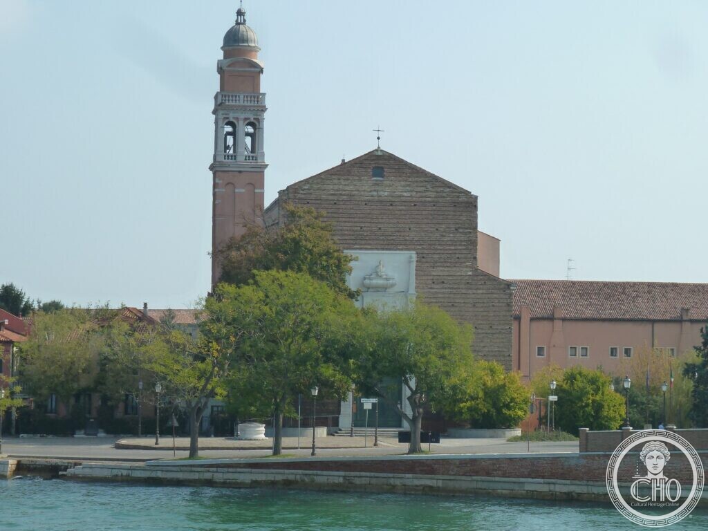 A plain brick church front with a white stone frame holding a marble urn, a tall bell tower topped by a small dome on the left, trees and a waterfront in front