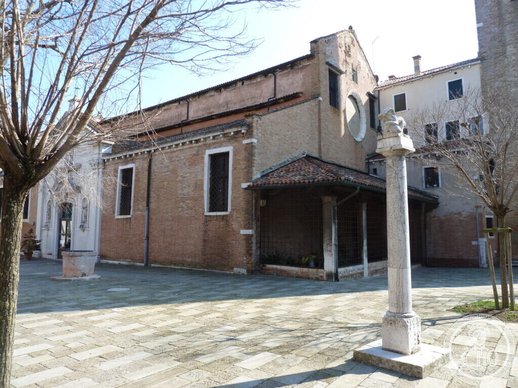 The long brick flank of a church with a low timber portico, a stone column topped by a worn stone lion in the foreground