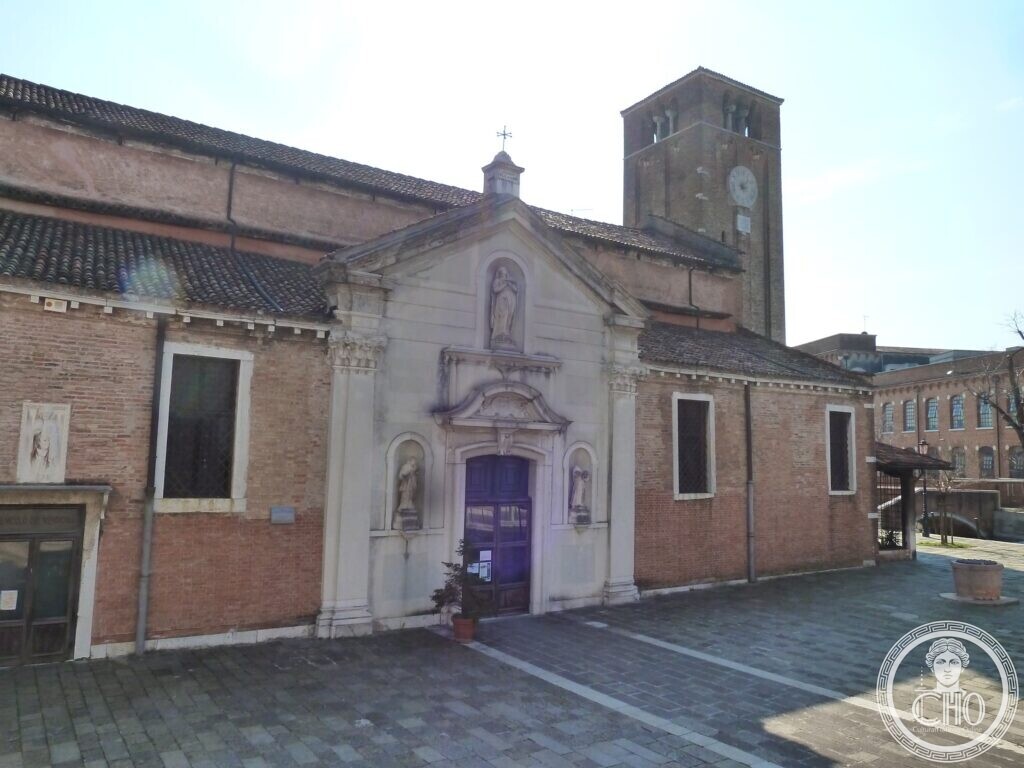 A small church with a white stone doorway set in a brick front, a square brick bell tower with a clock rising behind it, a paved campo in front