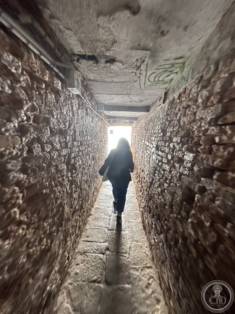 A narrow vaulted passage of bare brick with timber beams overhead, a walker silhouetted against the daylight at the far end