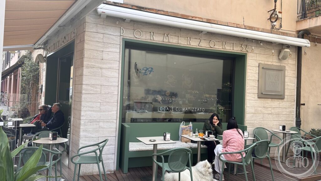 The words DON MINZONI 54 incised in wide-spaced Roman capitals in the travertine facing of a corner building, above a green-fronted café with outdoor tables