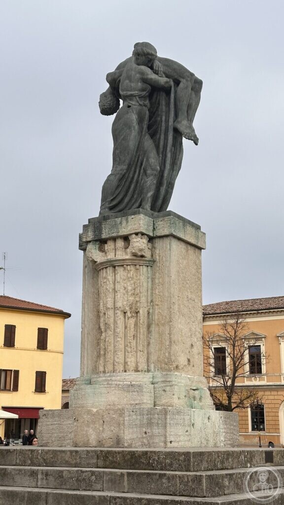 The bronze group of the war memorial at Santarcangelo di Romagna seen from the square: two draped women holding the body of a naked dead soldier, on a fluted stone stele