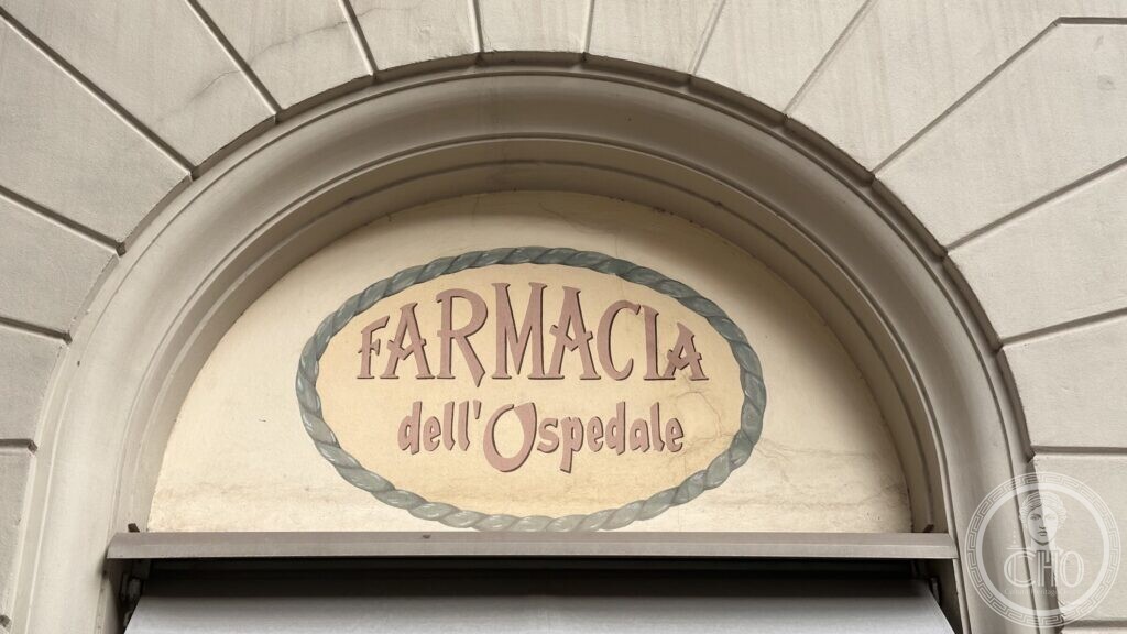 A painted oval sign in a frame moulded like twisted rope, reading FARMACIA dell Ospedale, set inside a stone arch over a doorway