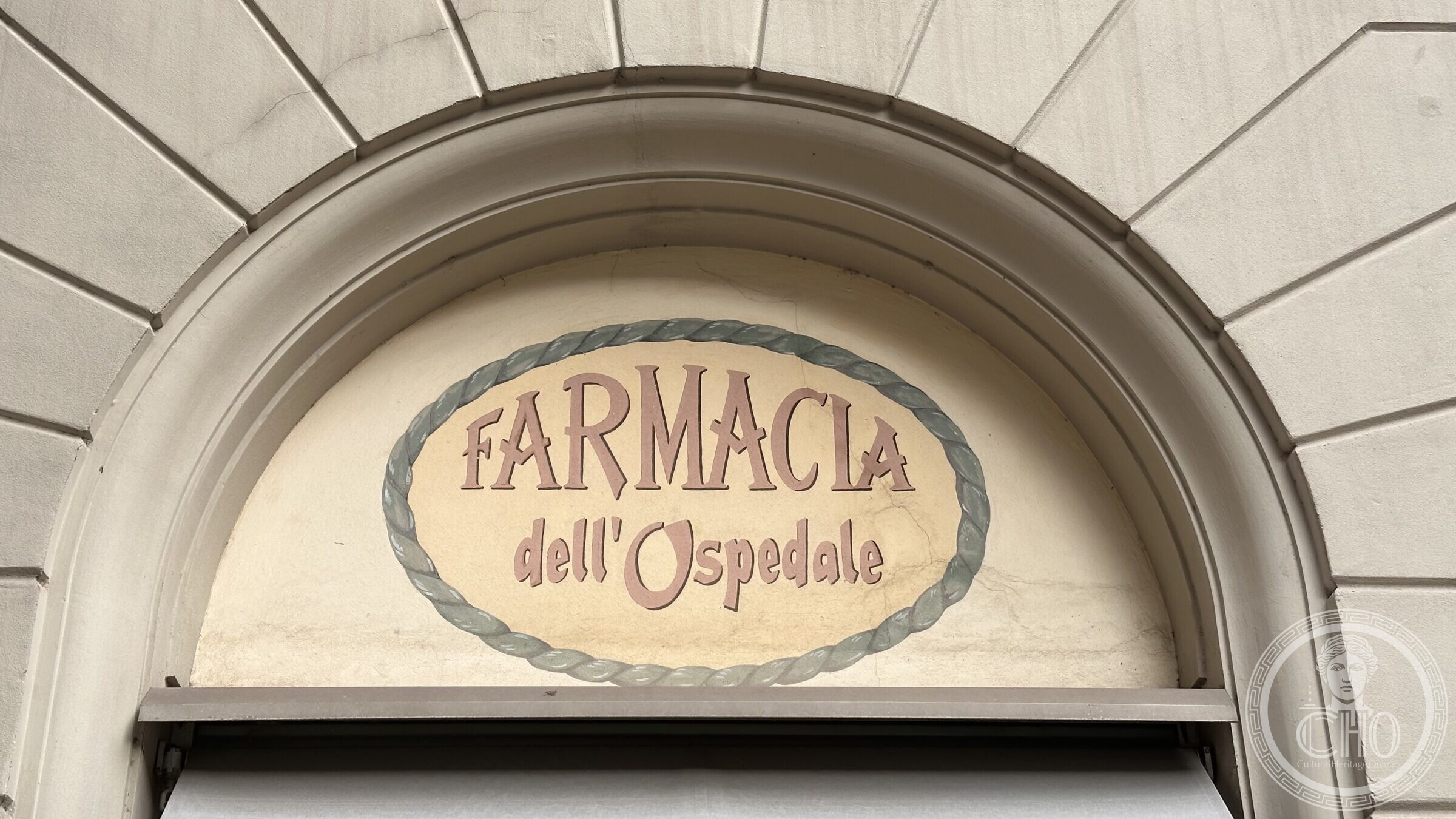 An oval painted sign reading FARMACIA dell'Ospedale, in a rope-moulded frame over a doorway