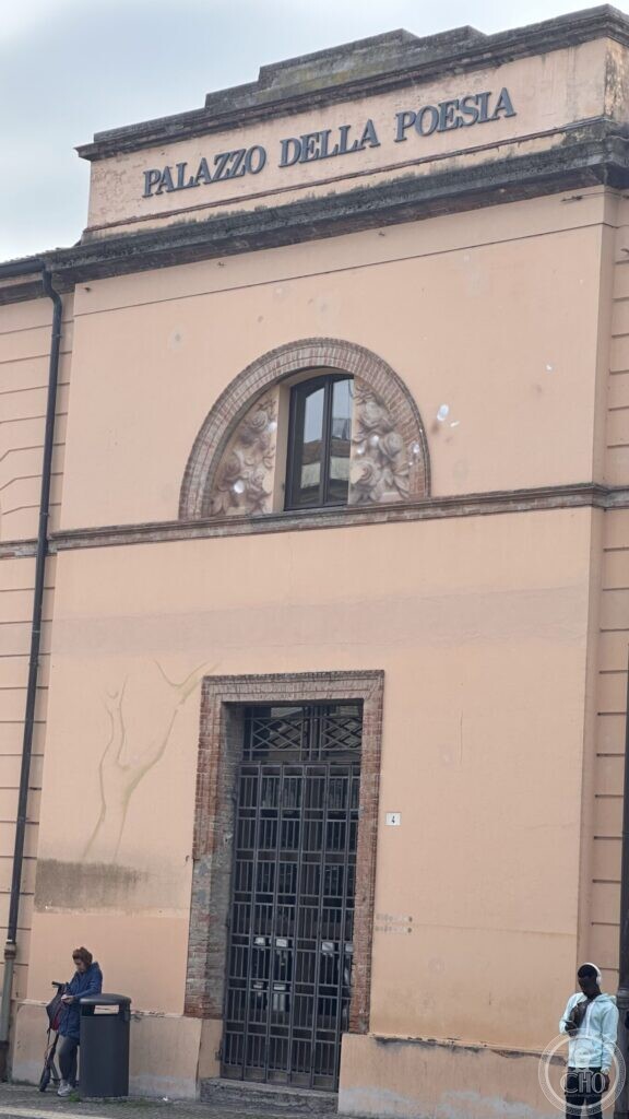 The front of the Palazzo della Poesia in Santarcangelo di Romagna: ochre render, a wide brick arch with a rose relief, an iron grille door numbered 4, and the name in metal letters on the attic